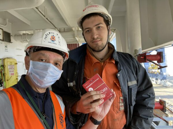 The Stella Maris chaplain on board ship handing a seafarer a mobile SIM card donated by Vodafone. Both are wearing protective clothing and hard hats.
