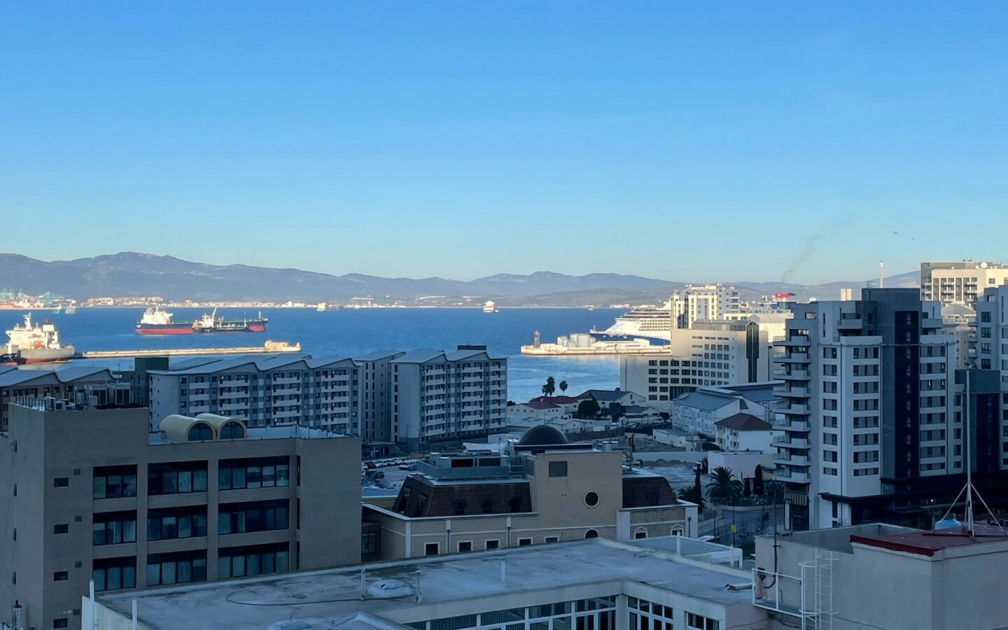 A view across apartments and buildings to the port of Gibraltar where a cruise ship is visible in the distance. This page also shows a video showing scenes from working life at sea. There are large ships at sea viewed from an aircraft or drone, an overhead view of a port, fishermen pulling in a large net, close up of a ship's controls, a close up of a man working on board ship wearing a blue hard hat