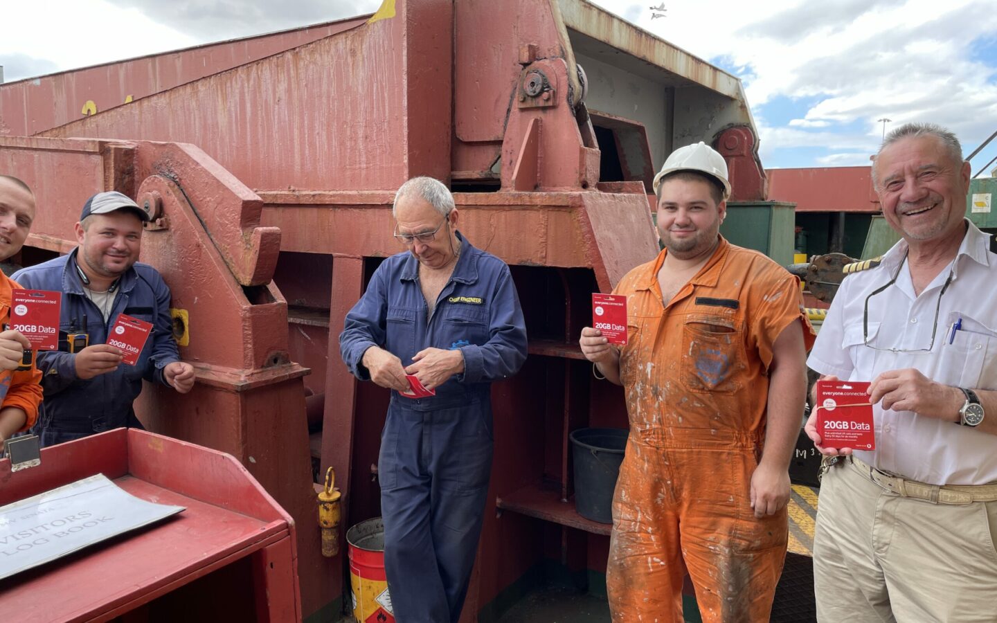 a group of five ship's crew members including the captain stood in front a large piece of equipment, smiling and holding SIM cards provided by Vodafone.