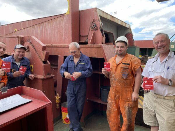 a group of five ship's crew members including the captain stood in front a large piece of equipment, smiling and holding SIM cards provided by Vodafone.